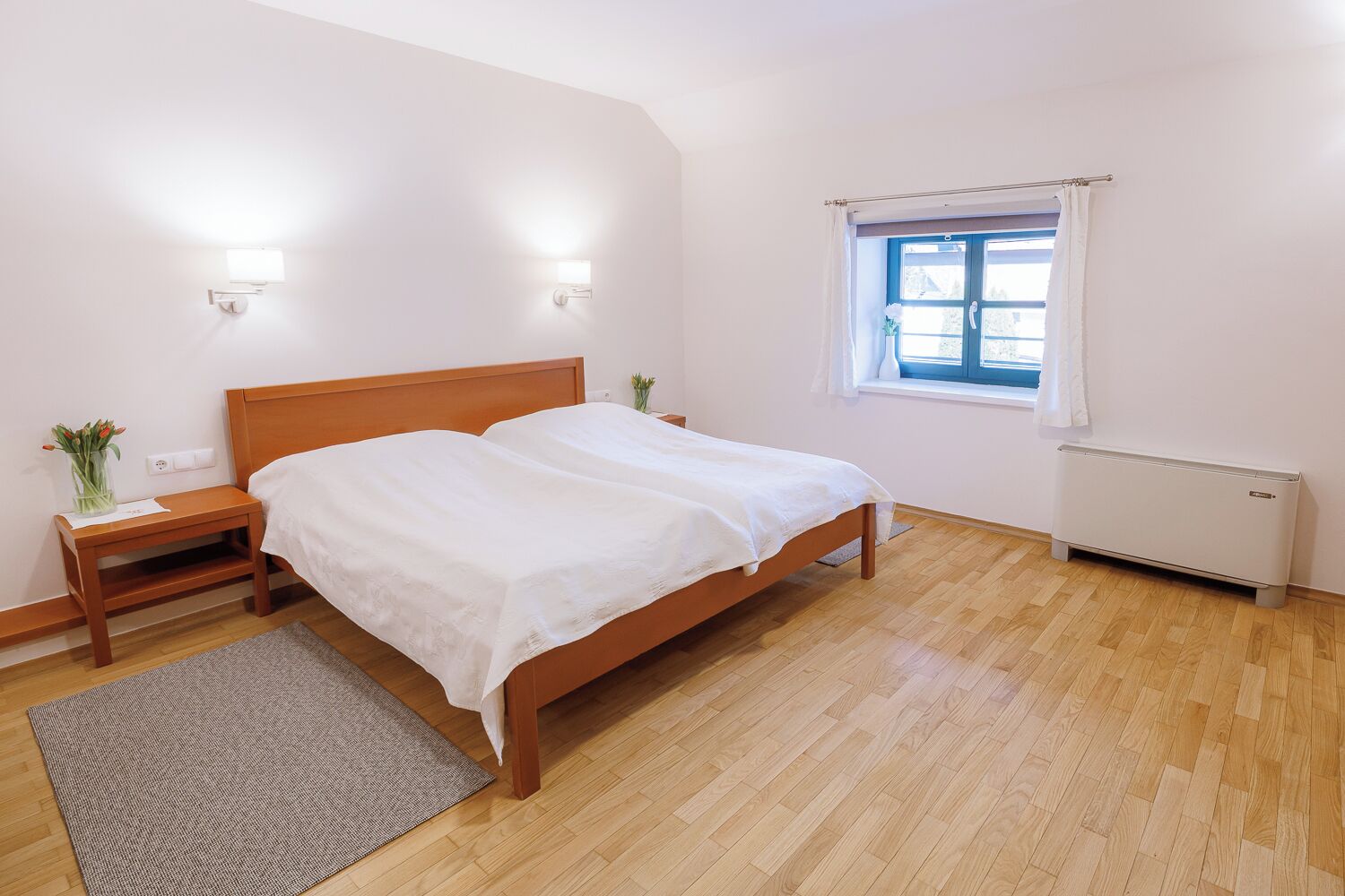 A bedroom with a wooden bed, bedside tables, a window with curtains, and a radiator.