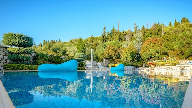 A clear blue pool with two blue floating structures, surrounded by lush greenery and a stone wall.