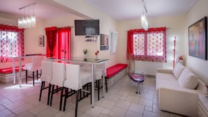 A dining area with a glass table, white chairs, and red curtains. There is a flat-screen TV mounted on the wall above the table.