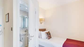 Habitación de hotel con cama, mesita de noche, lámpara fijada a la pared y baño visible a través de una puerta abierta.