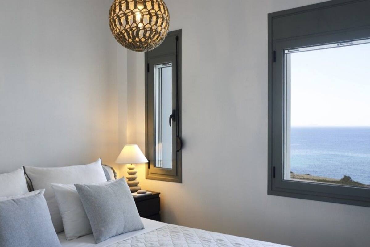 Ein Schlafzimmer mit Bett, Kissen, Nachttisch, Lampe, Spiegel und Fenster mit Meerblick.