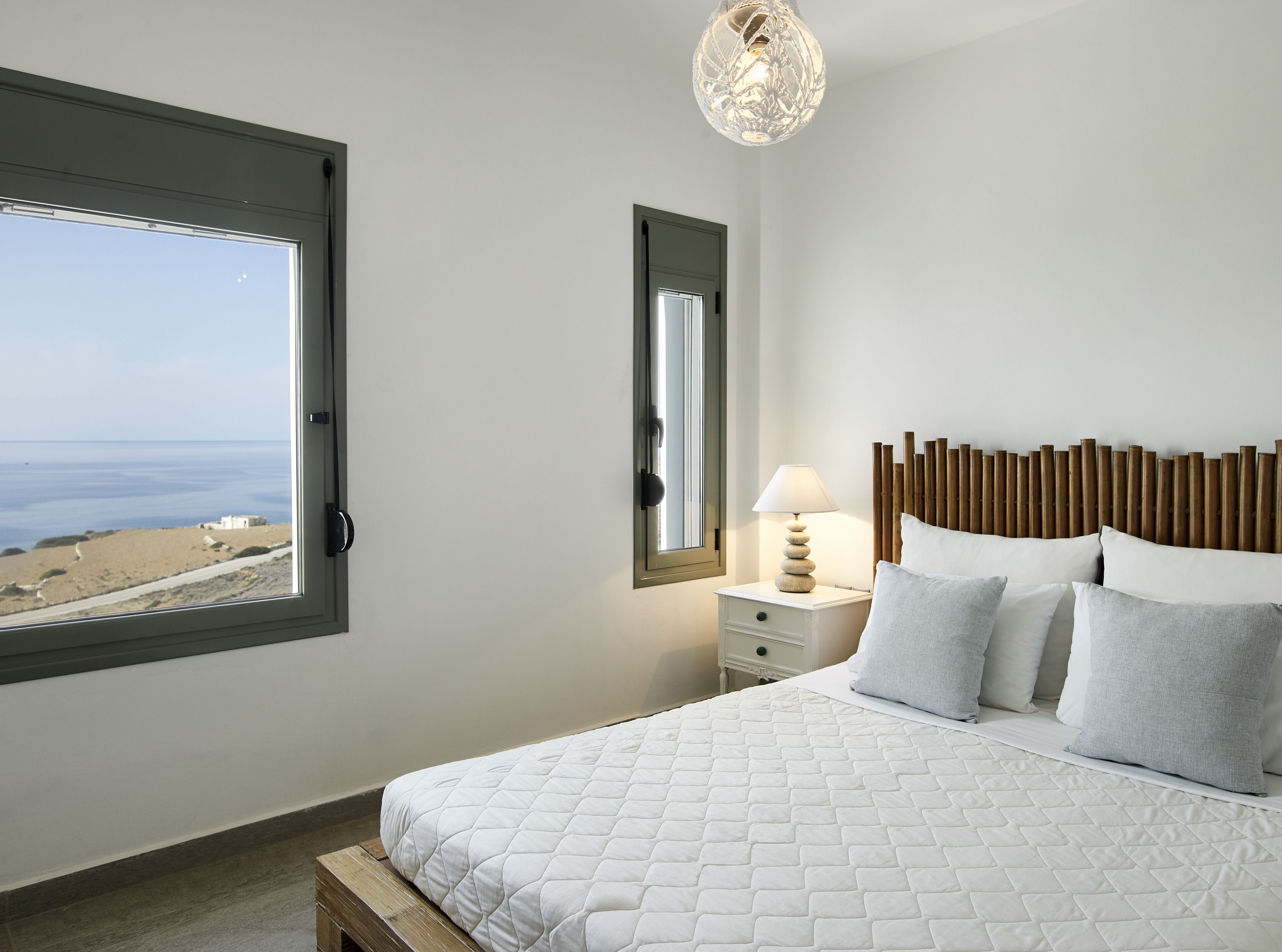 A bedroom with a large window, a bed with white bedding, a bedside table with a lamp, and a view of the sea.