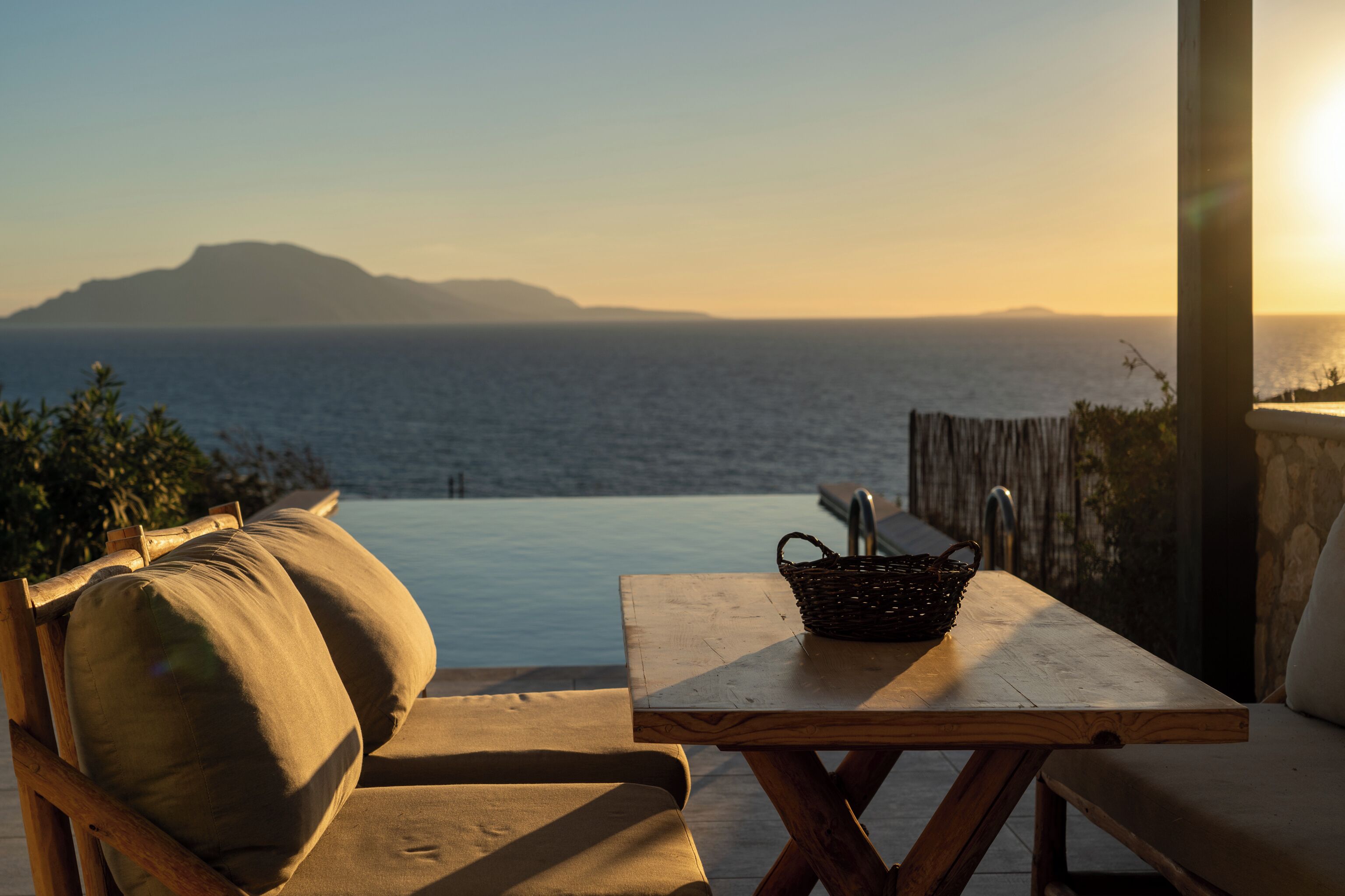 Ein Holztisch mit einem gewebten Korb, ein gepolsterter Stuhl und Blick auf das Meer bei Sonnenuntergang.
