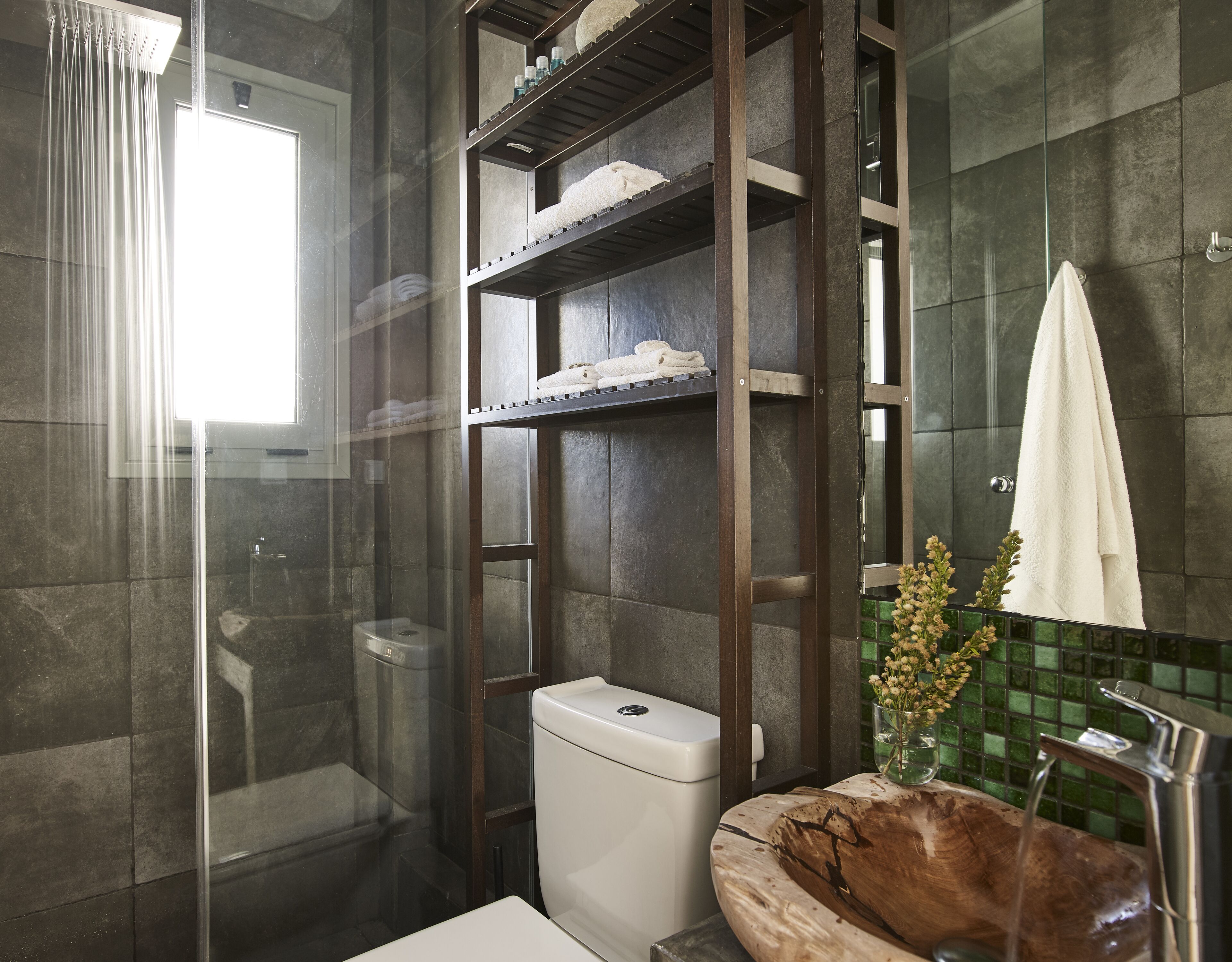 A modern bathroom with a glass shower enclosure, a wooden vanity with a bowl sink, and a shelf unit with towels.
