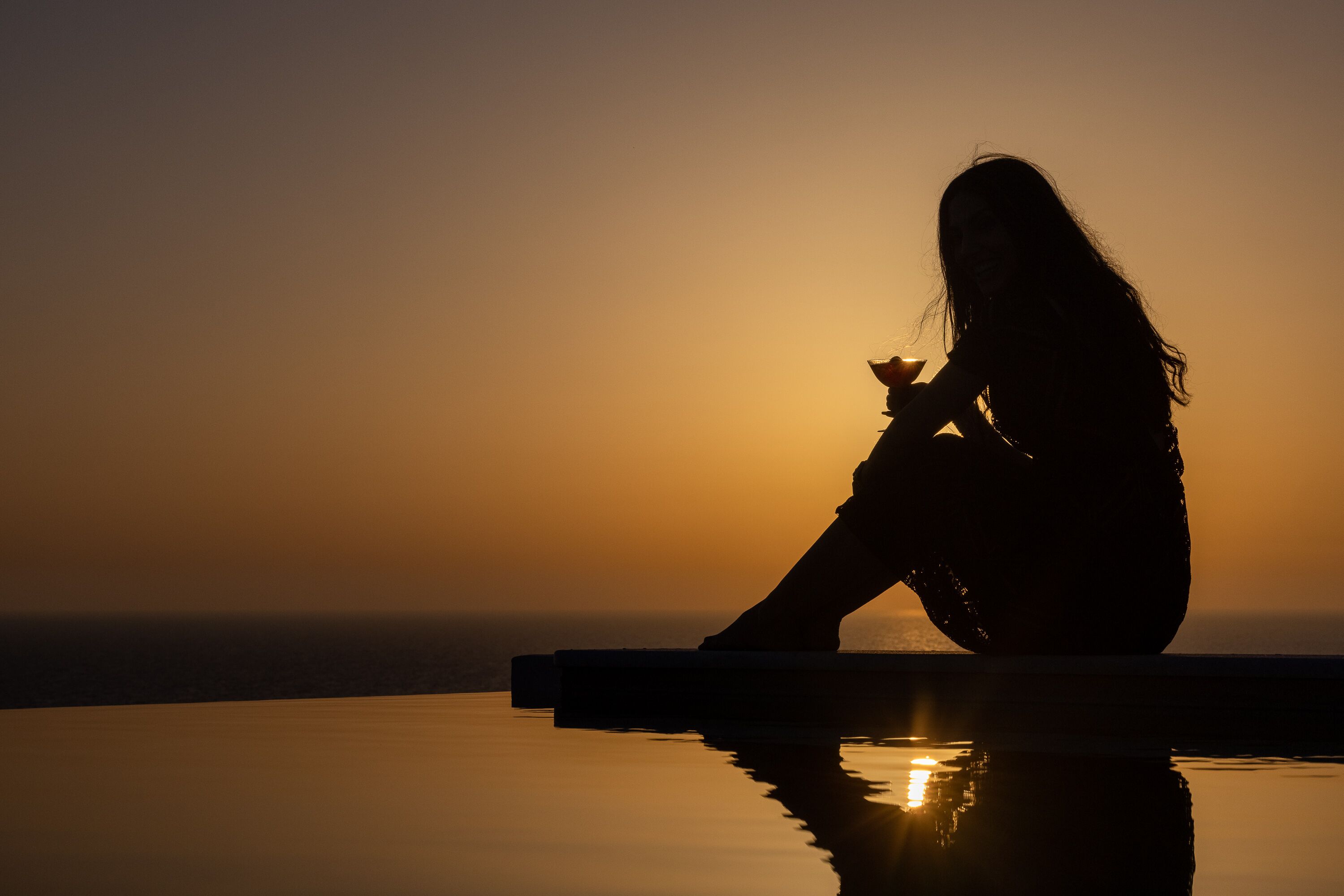 A silhouette of a person sitting by a reflective pool during sunset.