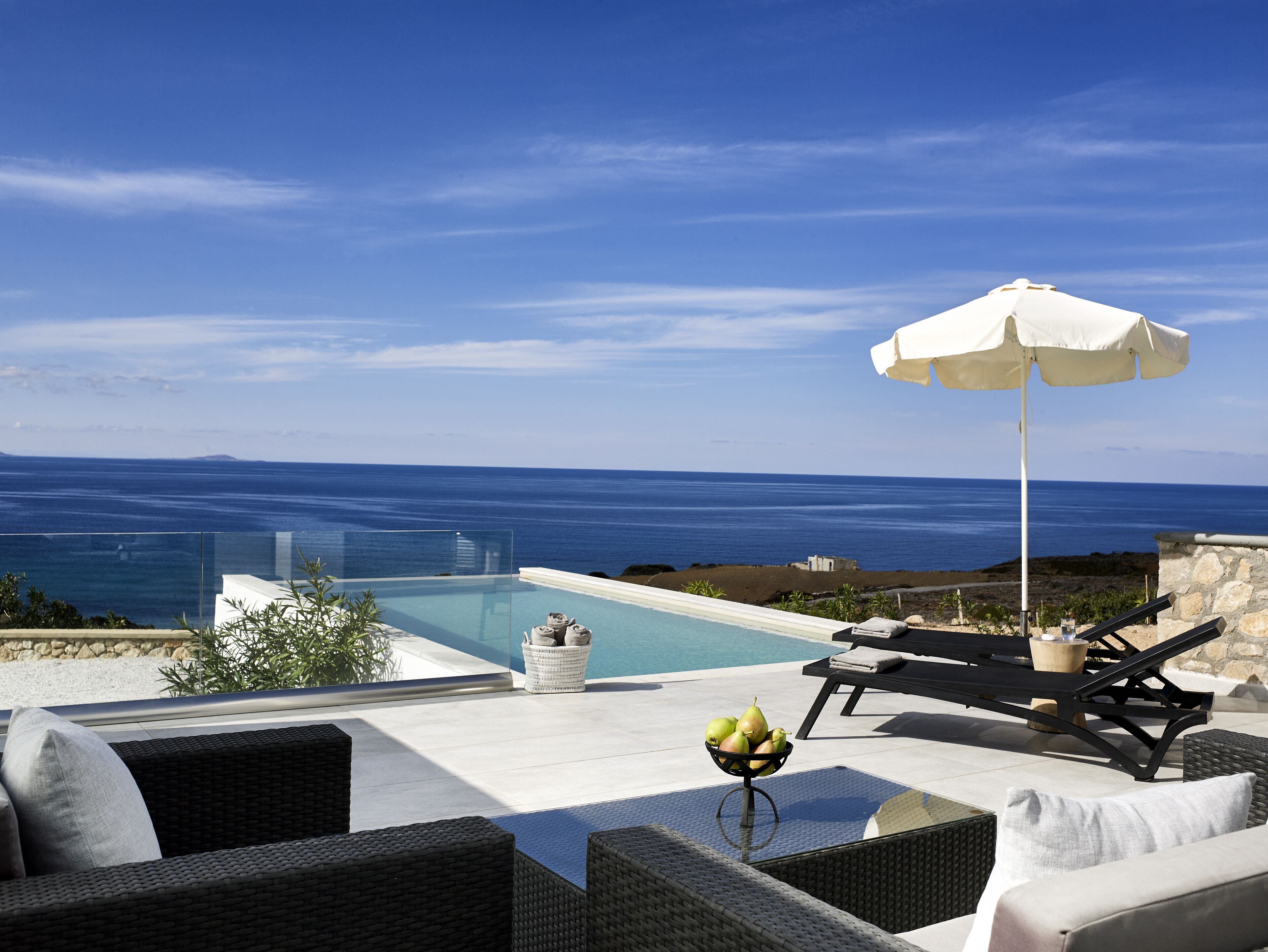A poolside area with a view of the sea, lounge chairs, an umbrella, and a fruit bowl.