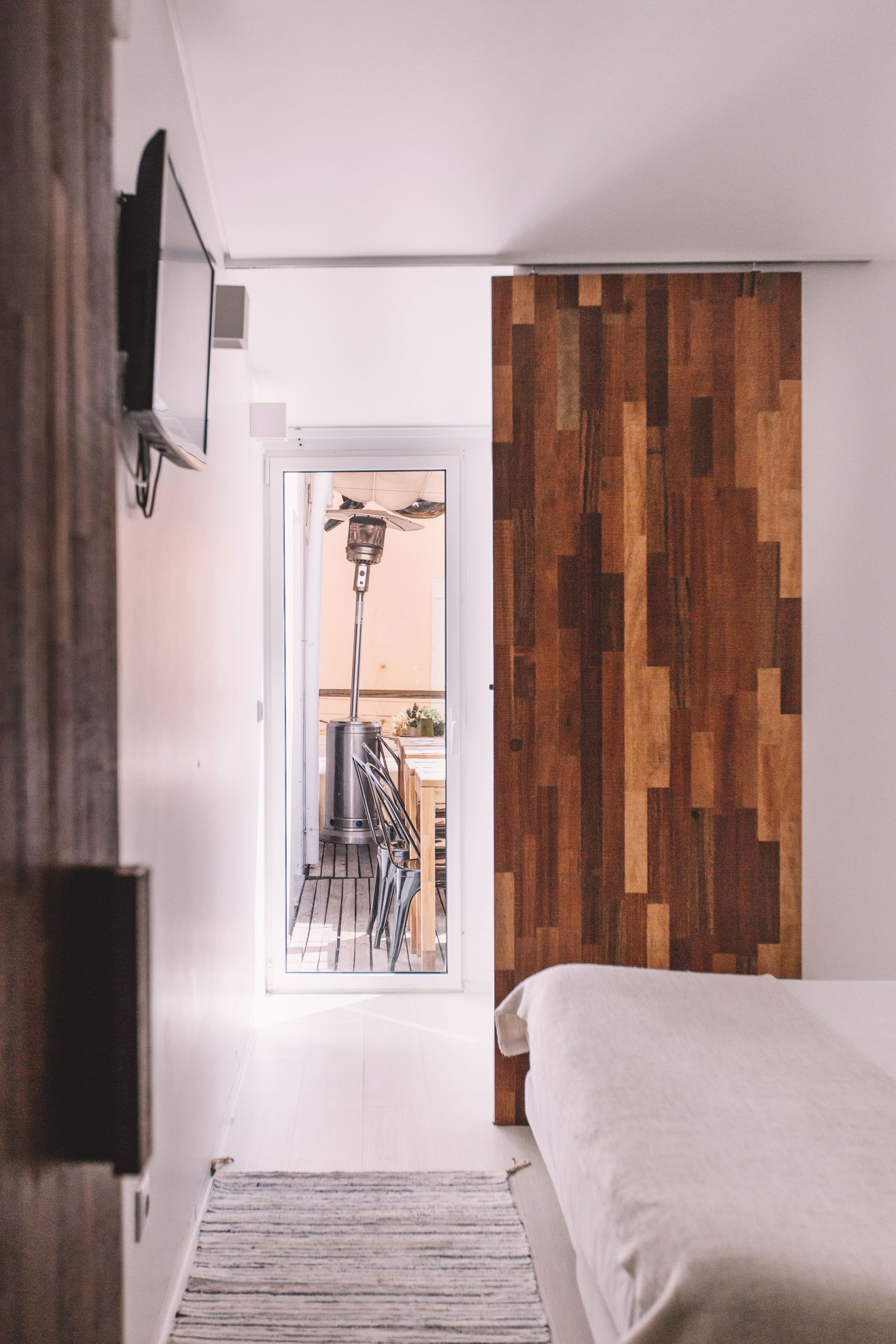 A bedroom with a wooden wall panel, a bed, a TV mounted on the wall, and a view of a balcony with outdoor furniture.
