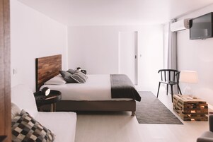 Quarto moderno com cama, mesa de cabeceira, abajur e televisão na parede.