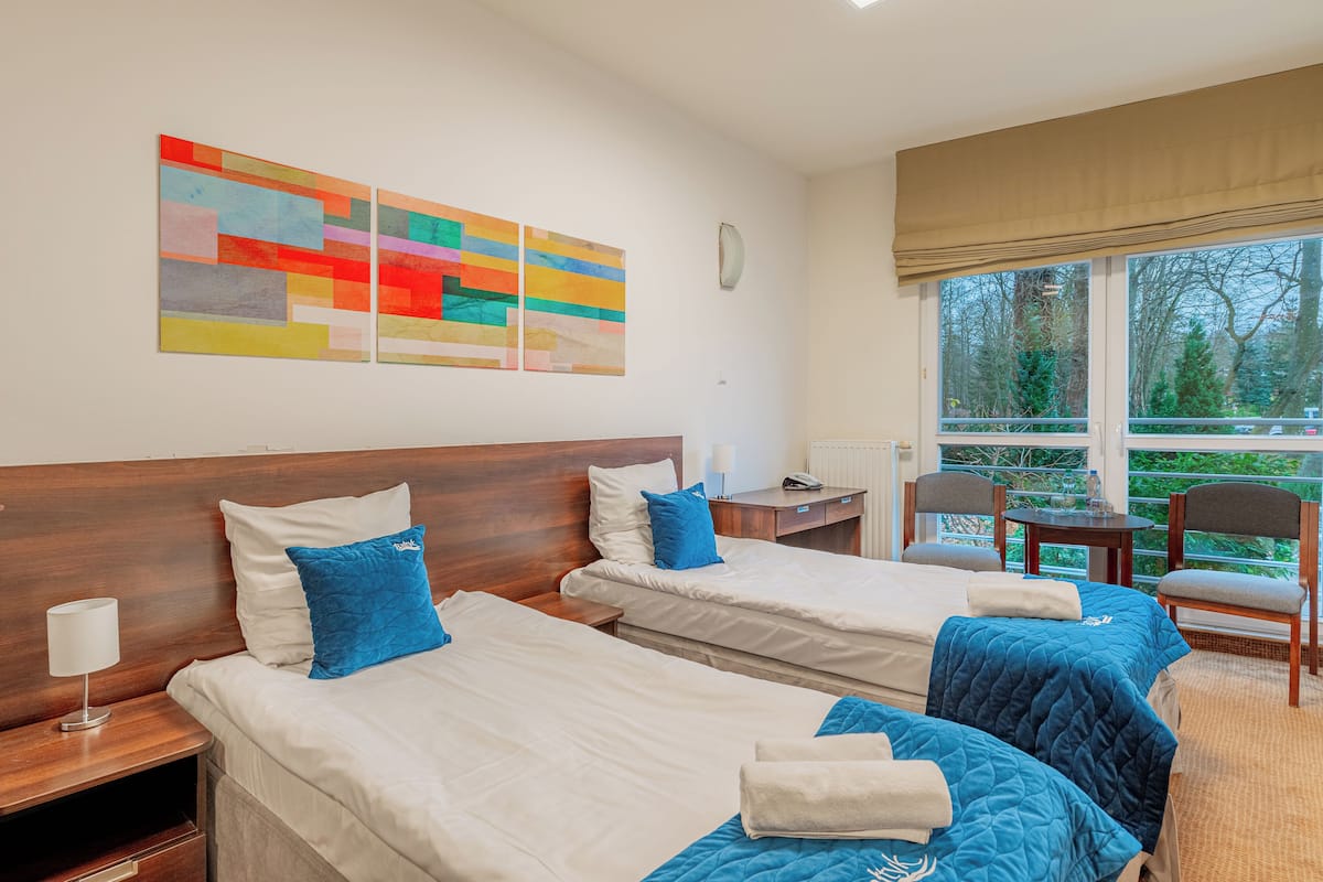 A hotel room with two beds, a window with a view of trees, a table with chairs, and a painting on the wall.