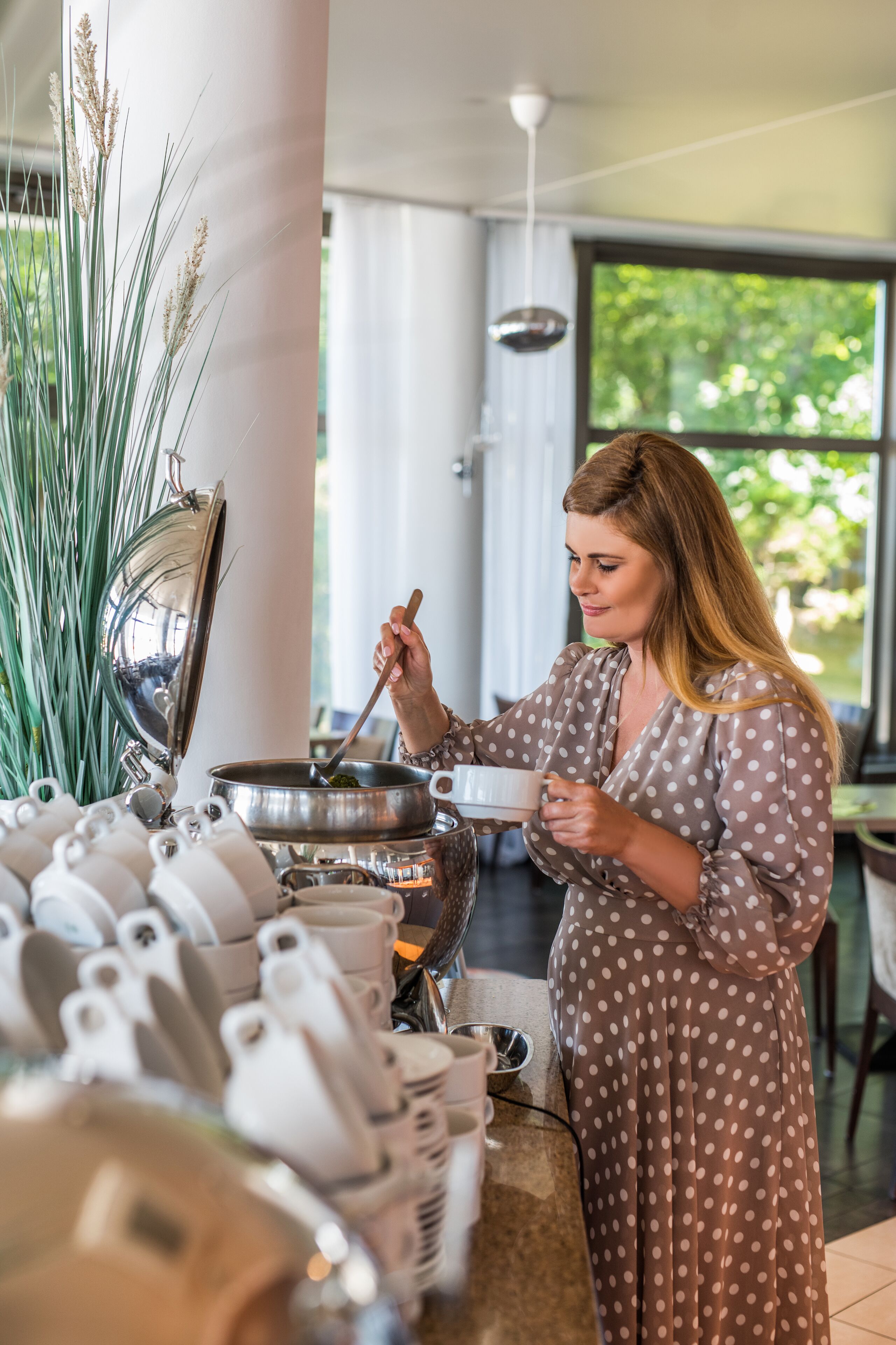 Une femme, vêtue d’une robe à pois, remue le contenu d’une marmite sur une cuisinière, tandis qu’une variété de théières et de tasses sont disposées au premier plan.