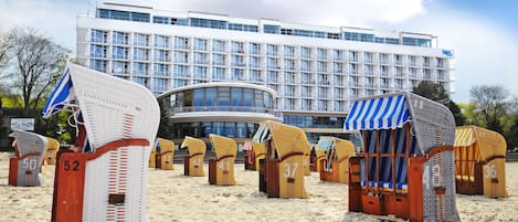 Un hotel de diseño moderno, con un edificio grande de múltiples ventanas y fachada de vidrio curvo. Frente al hotel, hay varias sillas de playa dispuestas sobre la arena.