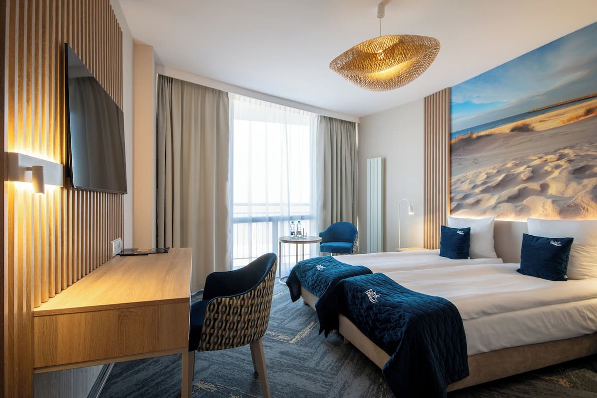 A hotel room with a bed, a desk, a chair, a small table, and a painting of a beach scene.