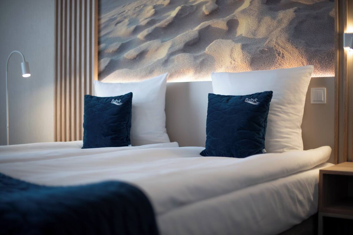 A neatly made bed with white and dark blue pillows, a wall-mounted light, and a wooden panel.