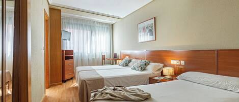 Una habitación de hotel con dos camas, un cabecero de madera, una mesita de noche con una lámpara, y un ventanal con cortinas.