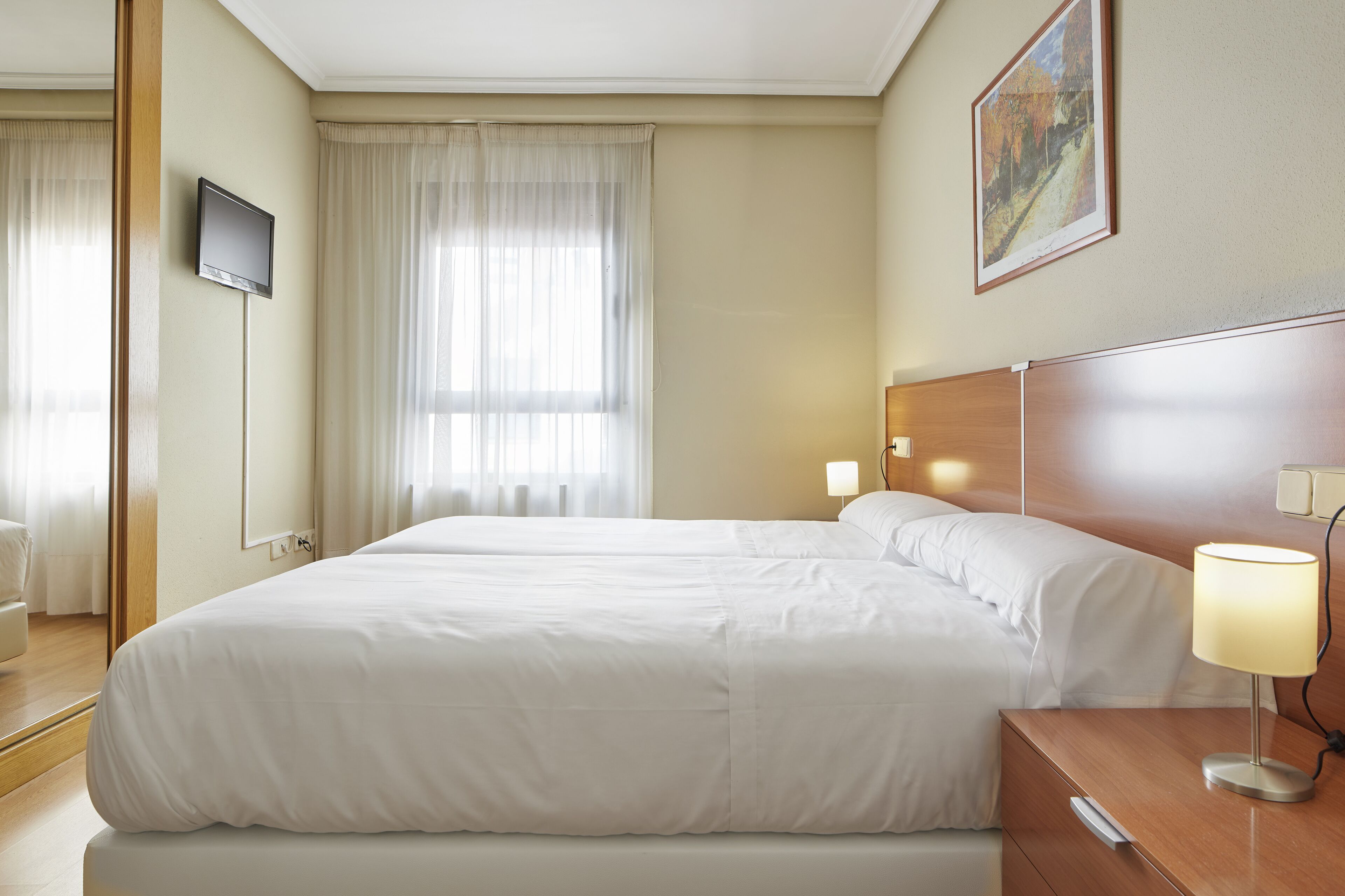 A hotel room with a large bed, a bedside table with a lamp, a TV mounted on the wall, and a painting on the wall.