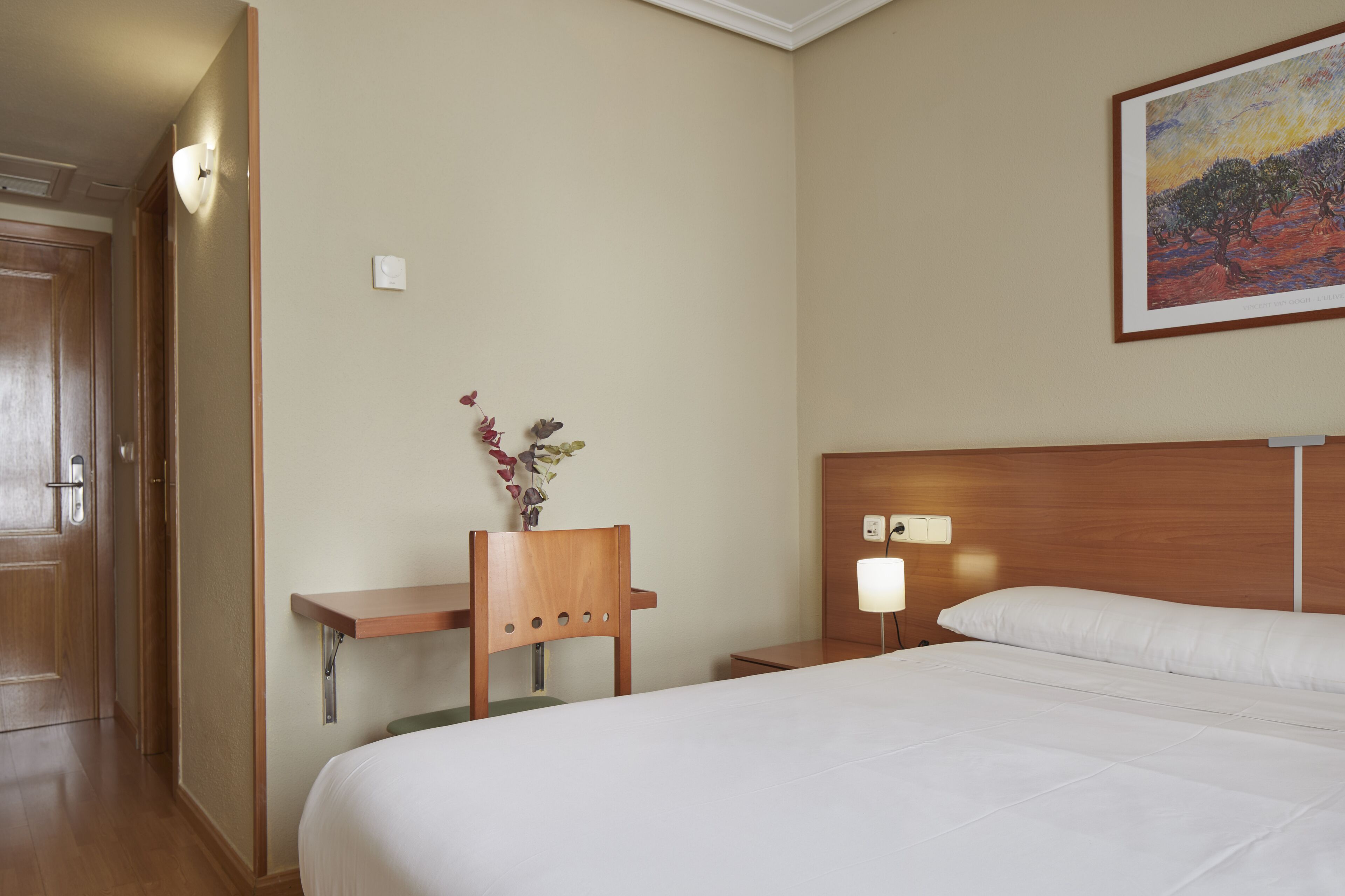 Ein Hotelzimmer mit einem Bett, einem Nachttisch, einer Wandlampe, einem Gemälde und einem kleinen Schreibtisch mit Blumen in einer Vase.