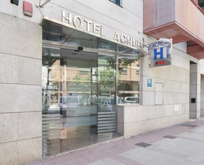 Entrada de un hotel con puerta de vidrio, detector de incendios y cartel con el nombre del hotel.