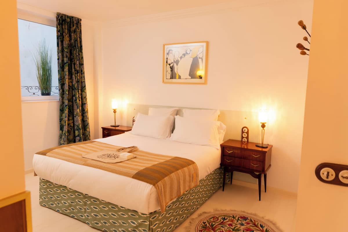A neatly made bed with a patterned bedspread, two bedside tables with lamps, a framed artwork on the wall, and a window with curtains.