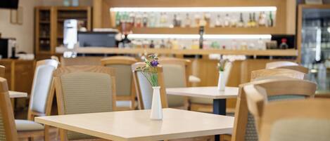 A restaurant with wooden tables and chairs, a bar area in the background, and a vase with flowers on each table.