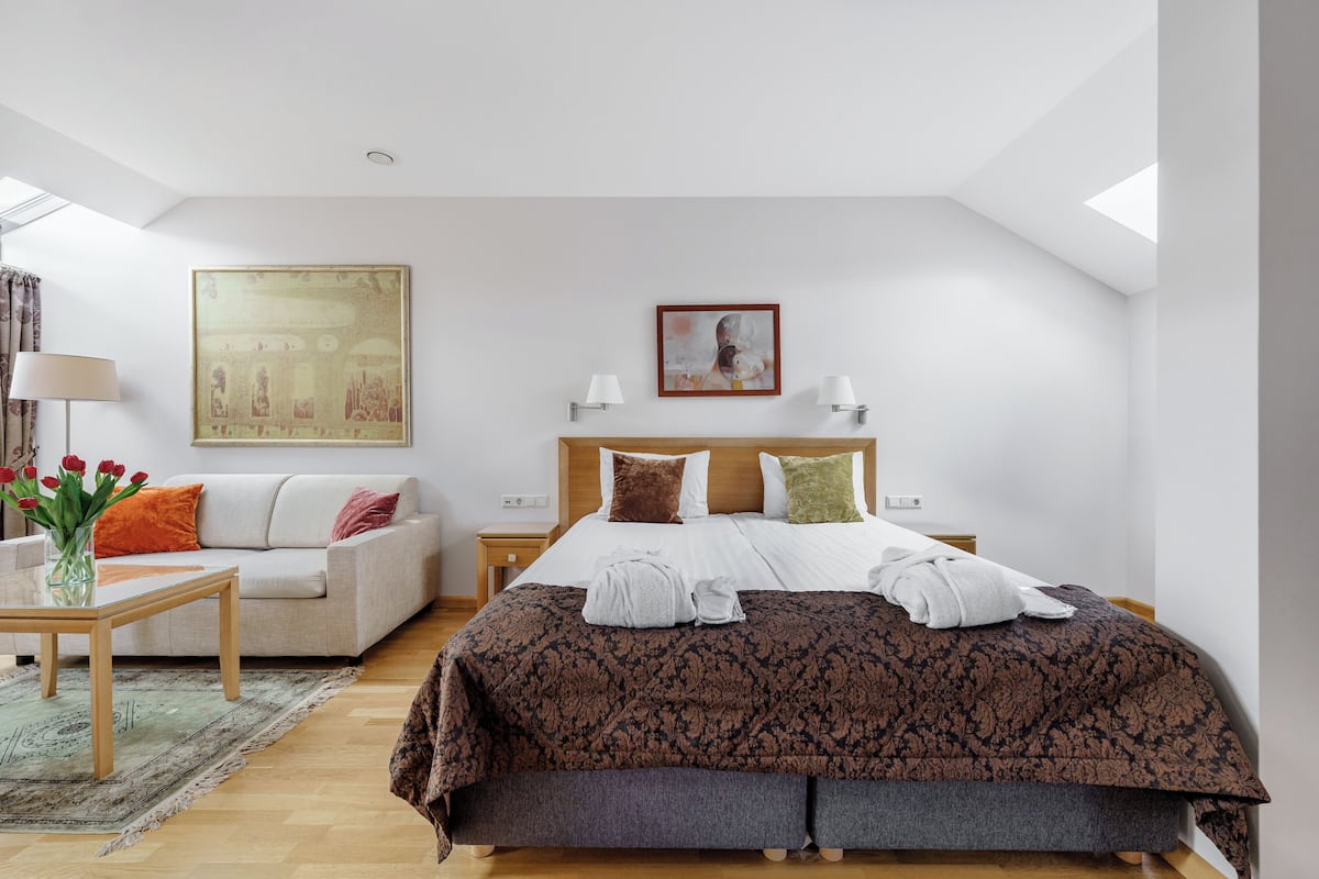 A modern hotel room with a large bed, a sofa, a small table, and a painting on the wall.