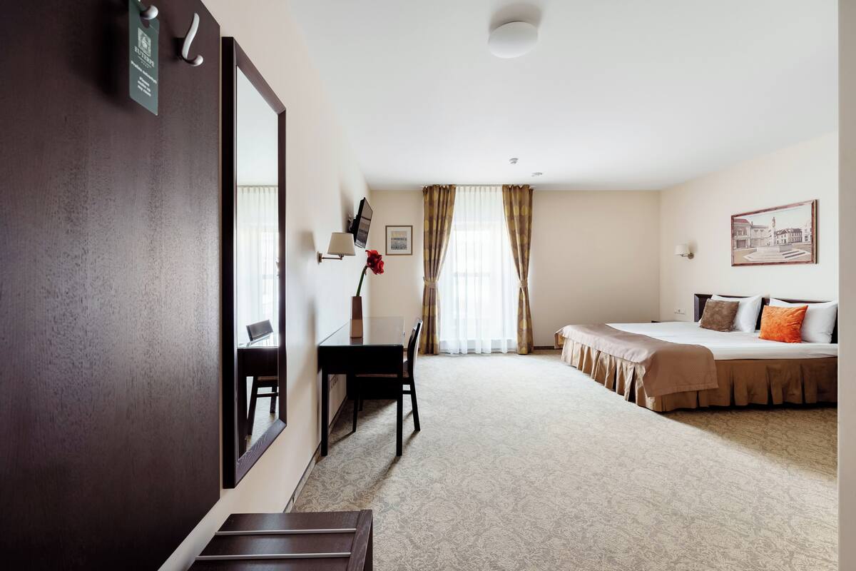 A hotel room with a bed, a desk, a chair, a mirror, a window with curtains, and a framed picture on the wall.