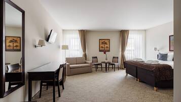 A hotel room with a bed, a sofa, a desk, and a chair. There are two windows with curtains, a lamp, and a painting on the wall.