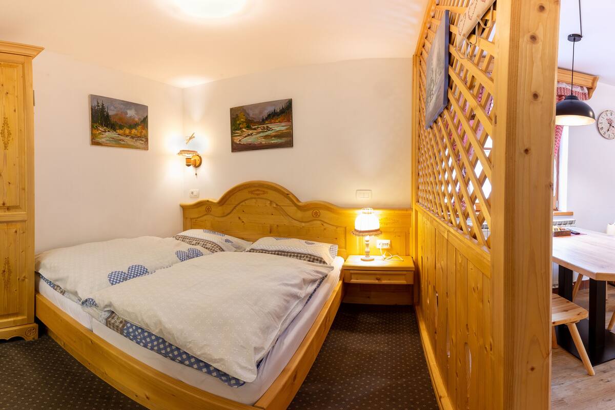 Una cama de madera con cabecera, dos cuadros en la pared, una mesita de noche con lámpara y un armario de madera.