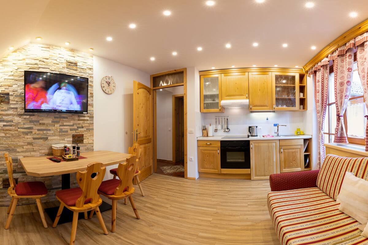 Una sala de estar acogedora con una mesa de comedor de madera y sillas, un televisor montado en una pared de piedra, y una cocina con armarios de madera y un ventanal con cortinas.