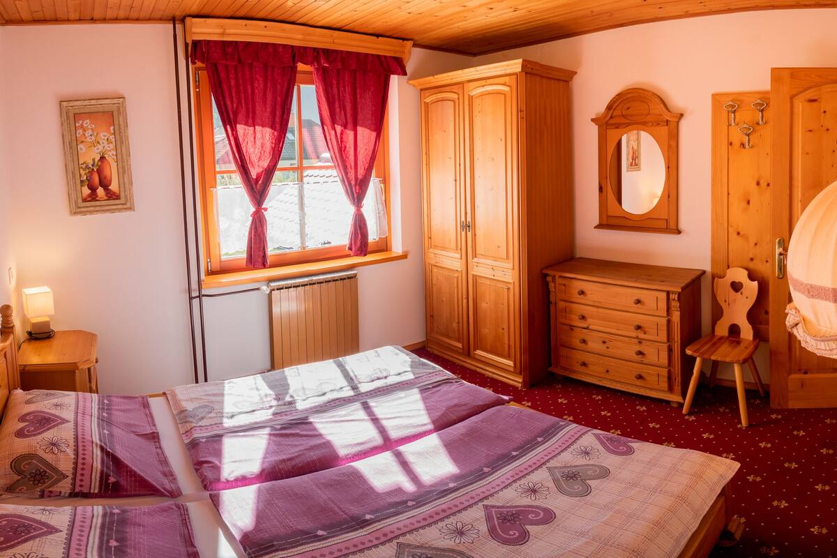 Un dormitorio con cama, armario de madera, cómoda, espejo, radiador y ventana con cortinas.