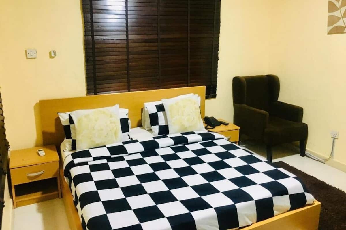 A bedroom with a checkered bedspread, a wooden headboard, a nightstand with a phone, a chair, and a window with blinds.