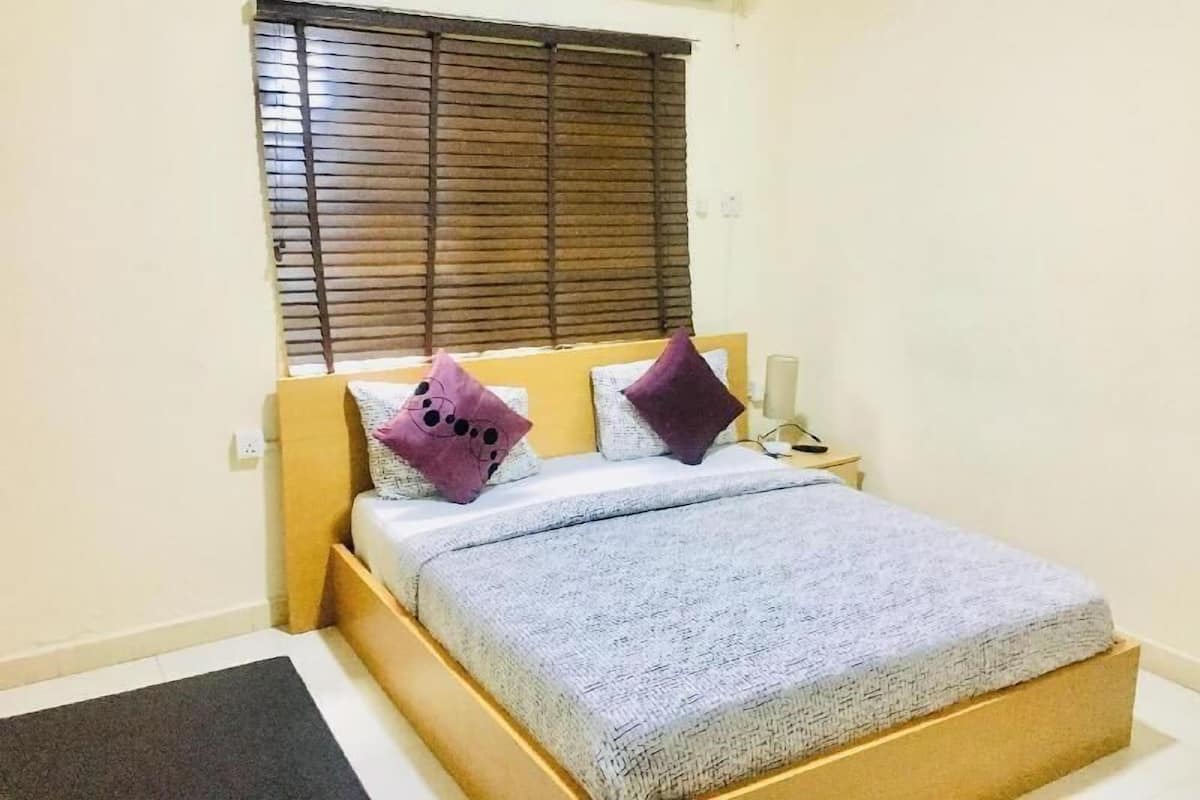 A single bed with a headboard, a bedside table with a lamp, and a window with blinds.