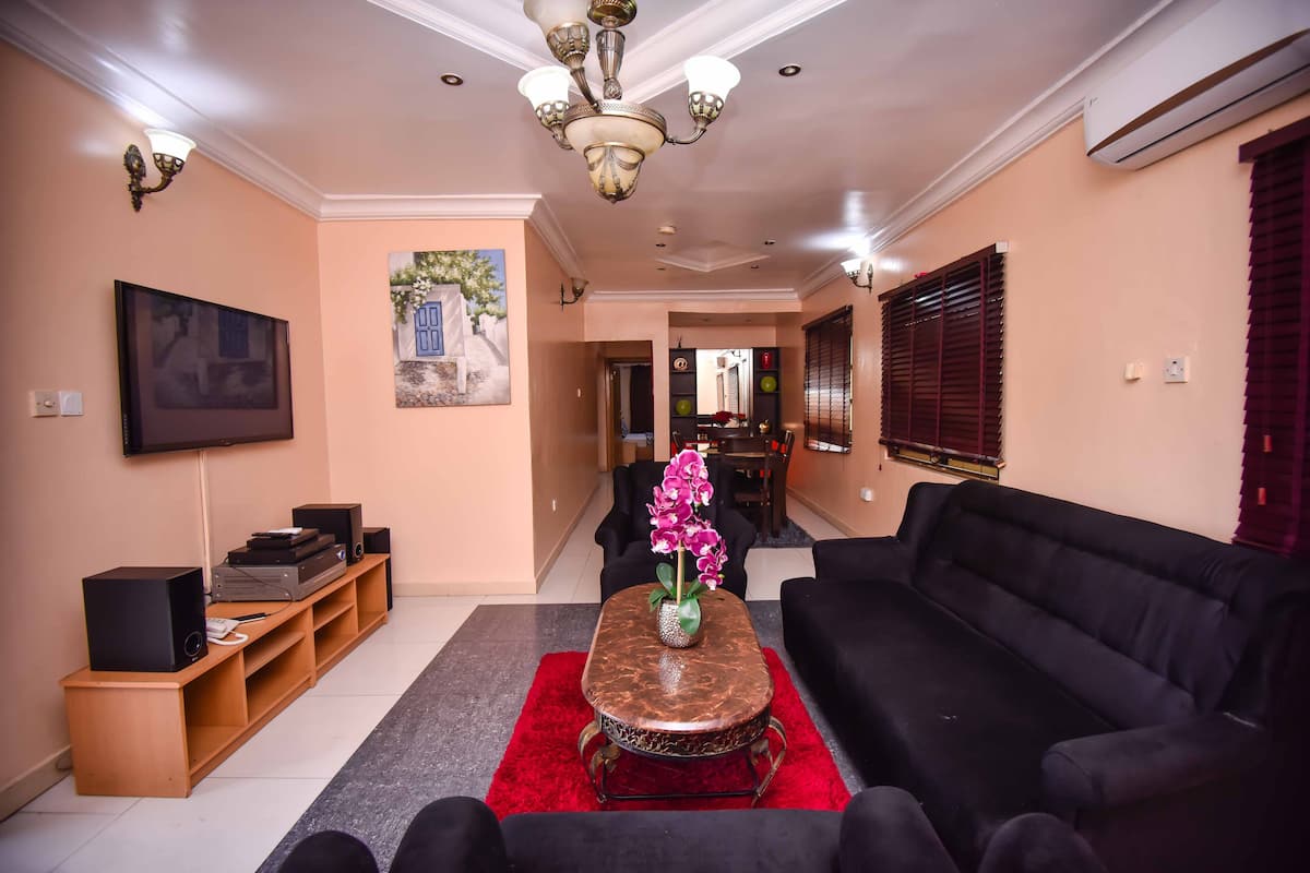 A living room with a black sofa, a red rug, a coffee table with a vase of flowers, a TV on a wooden stand, and a chandelier.