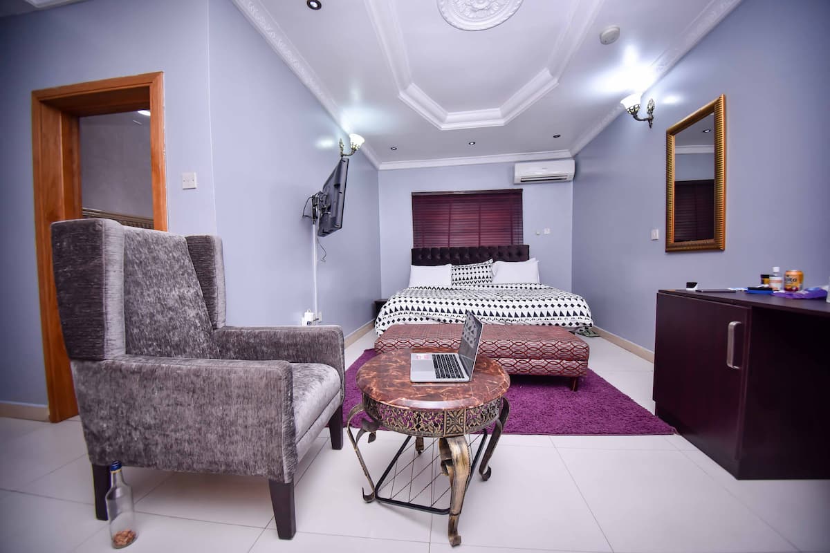 A bedroom with a bed, a desk with a laptop, a chair, a wardrobe, and a television.