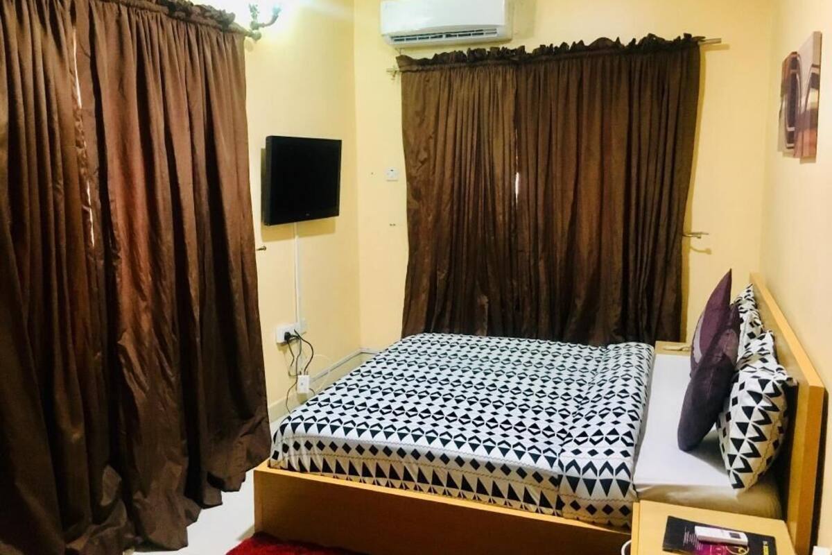A bedroom with a bed, curtains, an air conditioner, a TV, and a bedside table.