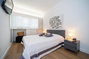 A neatly arranged bedroom with a bed, bedside tables, a wall-mounted TV, and a window with blinds. - Scoglietto 50 (Castiglione della Pescaia)