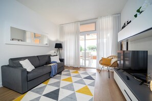 A modern living room with a grey sofa, a patterned rug, a television, and a sliding glass door leading to an outdoor area. - Scoglietto 50 (Castiglione della Pescaia)