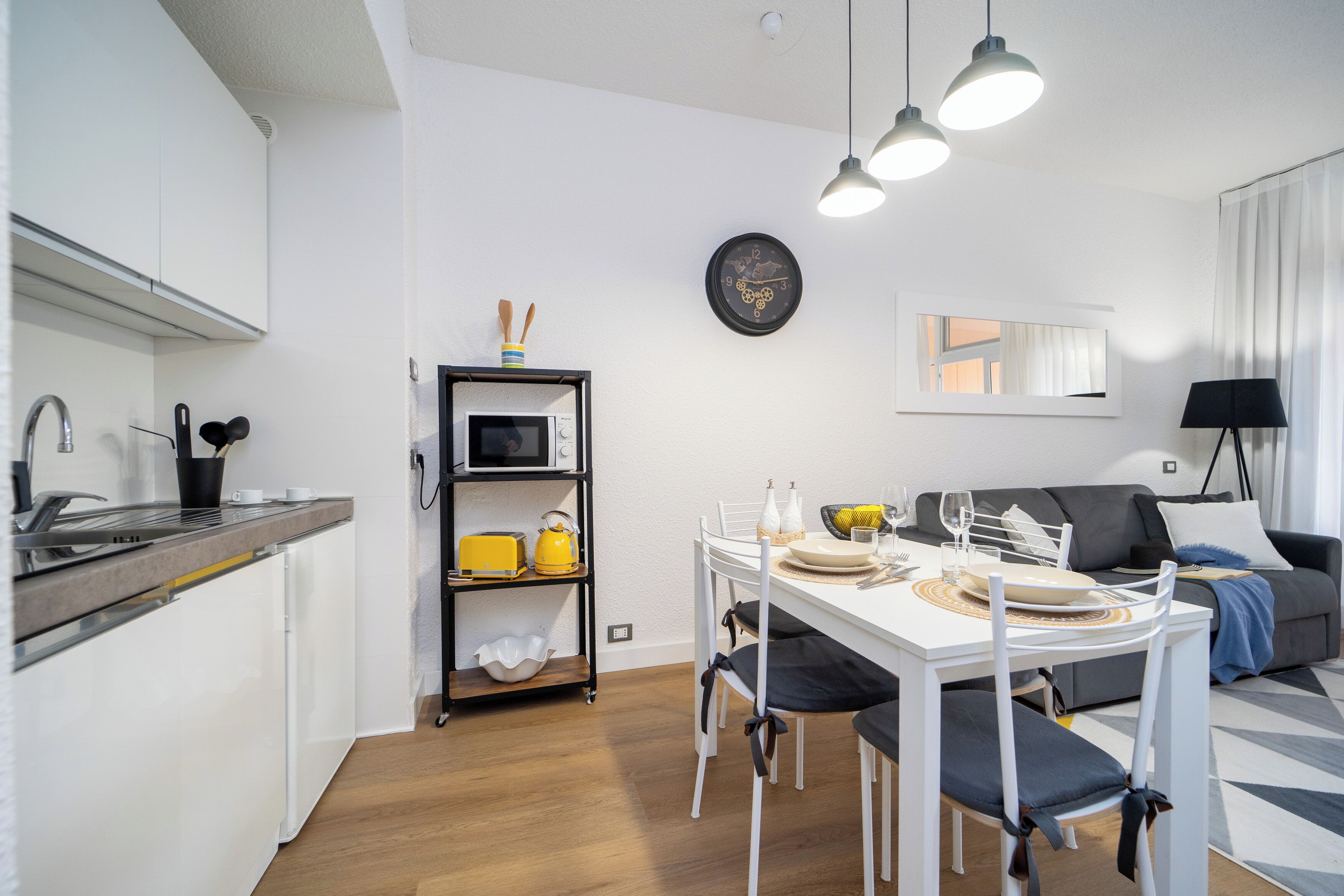 Una cocina moderna con armarios blancos, un microondas y una estantería con utensilios de cocina. Hay una zona de comedor con una mesa blanca, sillas y un sofá negro. Un reloj y un espejo están en la pared.