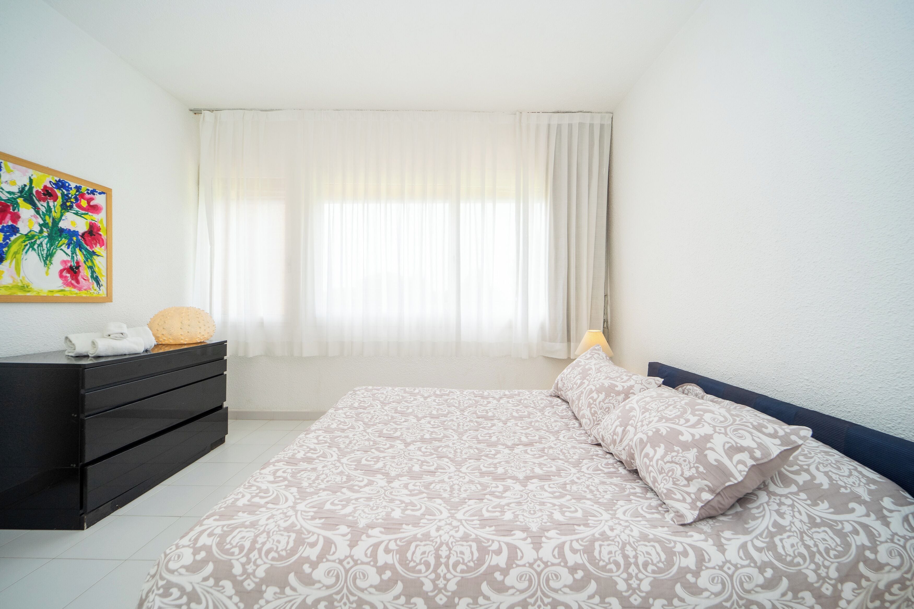 A bedroom with a bed, a dresser, a painting, and a window with curtains.