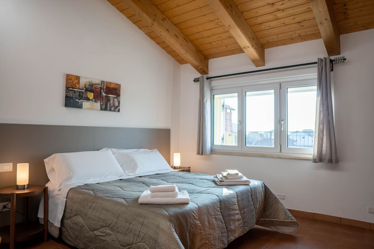 A neatly made bed with a gray quilt, a painting on the wall, and a window with curtains.