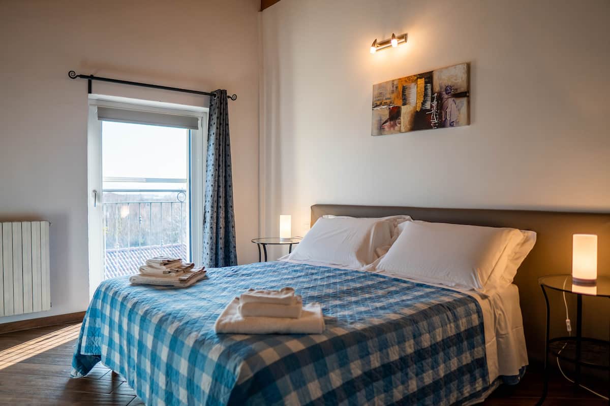 A neatly made bed with a checkered bedspread, a bedside table with a lamp, a window with curtains, and a painting on the wall.