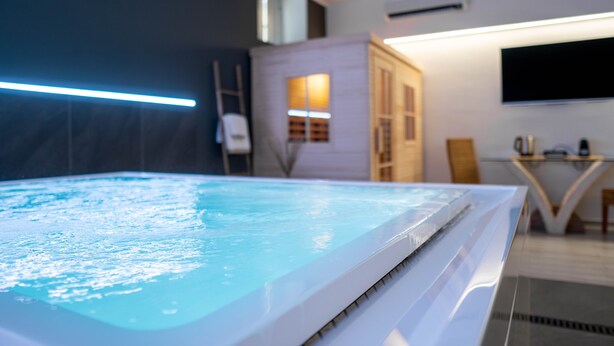 Una piscina interior moderna con superficie de agua cristalina, borde blanco y mampara de vidrio. Se observa un televisor montado en la pared y una pequeña mesa con una silla al fondo.