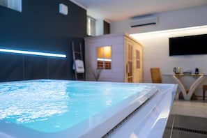 A modern indoor swimming pool with a clear water surface, a white edge, and a glass partition. There is a television mounted on the wall and a small table with a chair in the background. - Residence Le Palme (Torrazza Piemonte)
