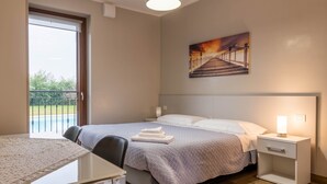 Habitación de hotel con cama, mesita de noche, mesa y silla. Hay un cuadro en la pared y vistas a un balcón con barandilla.