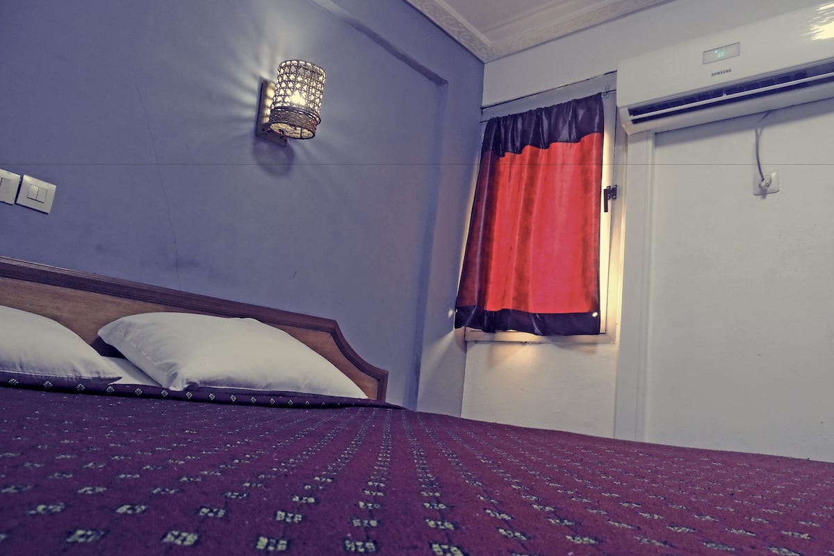 A bedroom with a bed, a wall-mounted light fixture, a window with a red curtain, and an air conditioning unit.