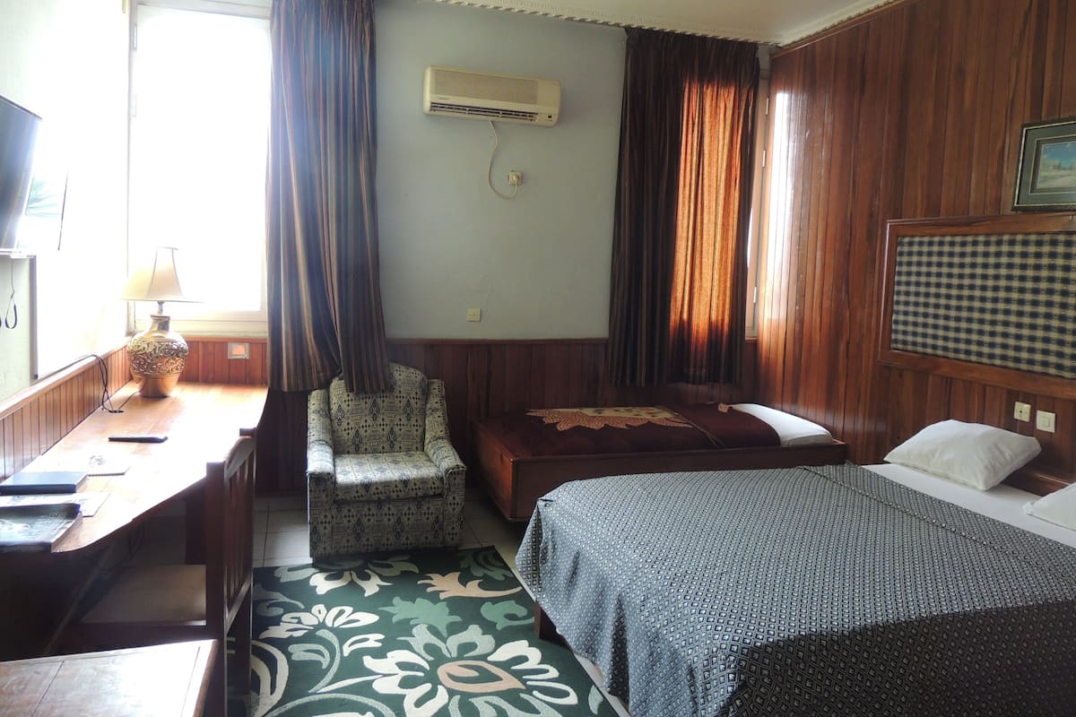A hotel room with a bed, a chair, a desk, and an air conditioning unit.