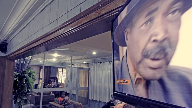 A living room with a large flat-screen TV, a sofa, a coffee table, and a potted plant. - Saffana Hôtel (Douala)
