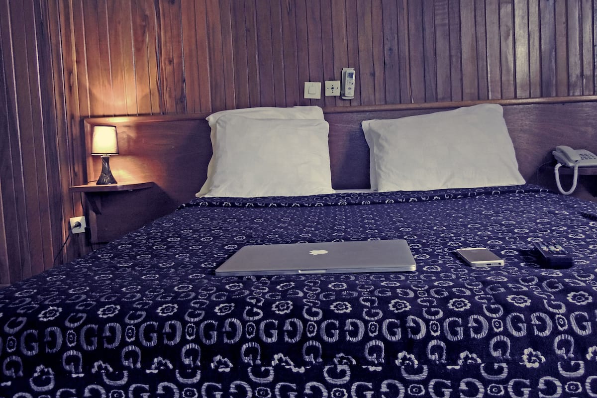 A hotel room with a wooden headboard, a bed with a patterned bedspread, a laptop, a phone, and a bedside table with a lamp.