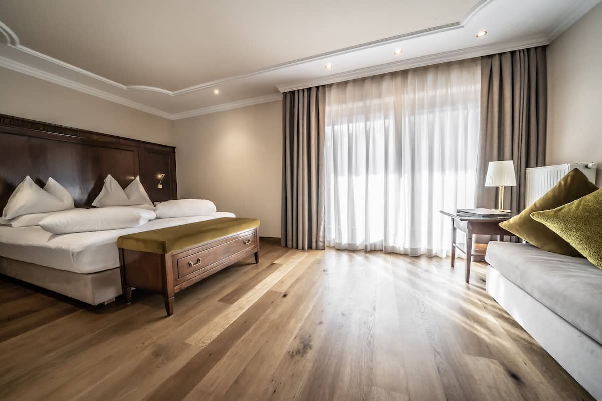 Habitación de hotel con piso de madera, una cama grande con ropa blanca, una mesita de noche de madera, una pequeña mesa con una lámpara y un sofá con un cojín verde.