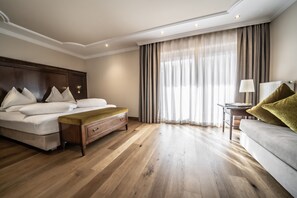 A hotel room with a wooden floor, a large bed with white linens, a wooden nightstand, a small table with a lamp, and a sofa with a green throw pillow. - Hotel Luna Mondschein (Ortisei)