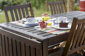 Une table en bois, dressée pour le petit-déjeuner, avec un verre de jus d’orange, une tasse de café, une assiette contenant une part de gâteau et un bol de fruits.